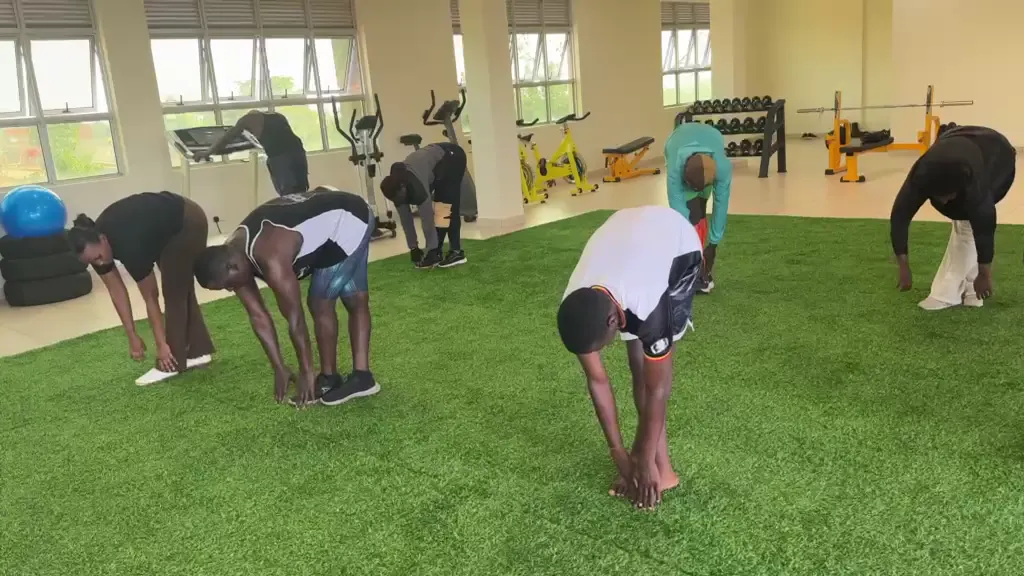 Members stretching on the turf in daylight, with cardio machines, spin bikes and the dumbbell rack behind them